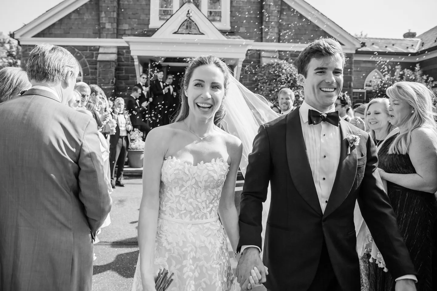 Black and white frame of the couple walking out of the chapel through smiling guests