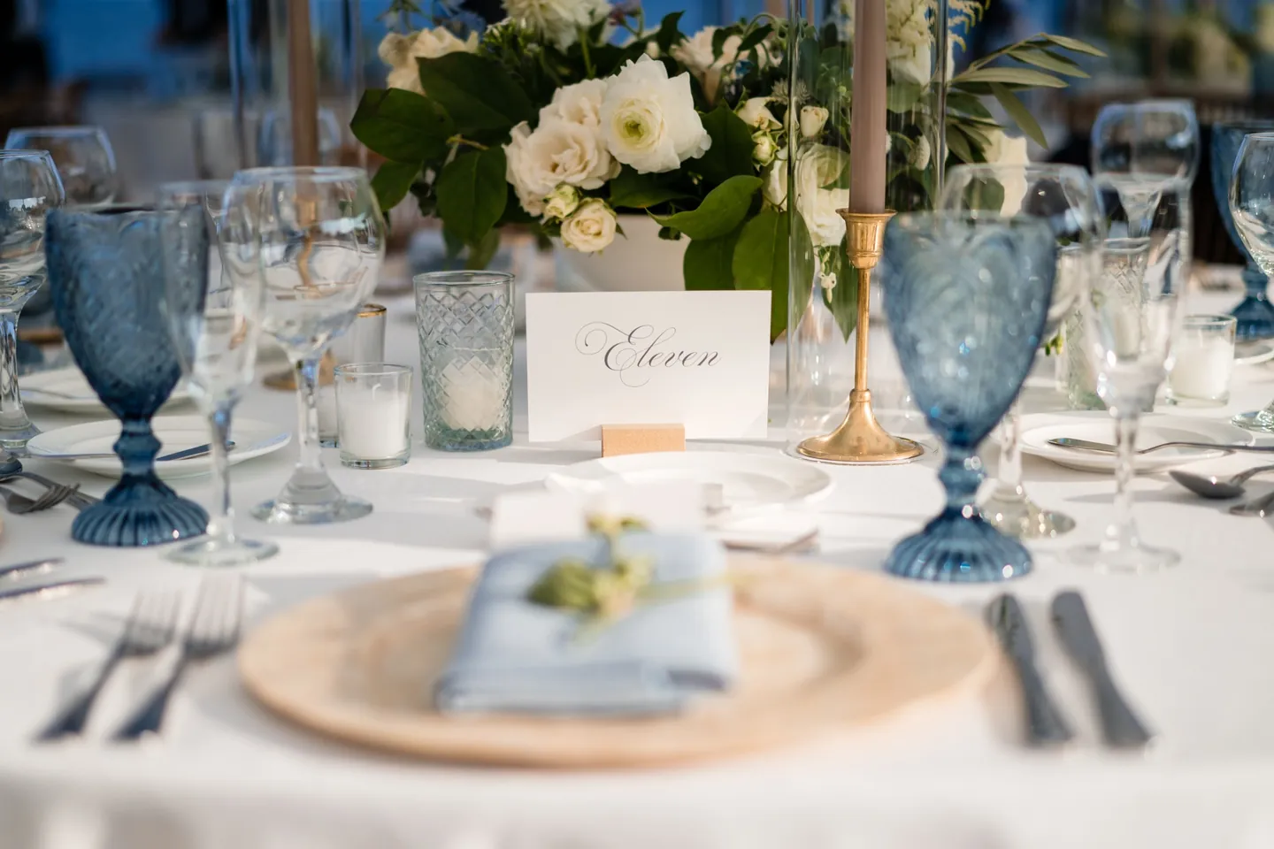 Blue glass goblets, a gold charger and a folded blue napkin under white hydrangea