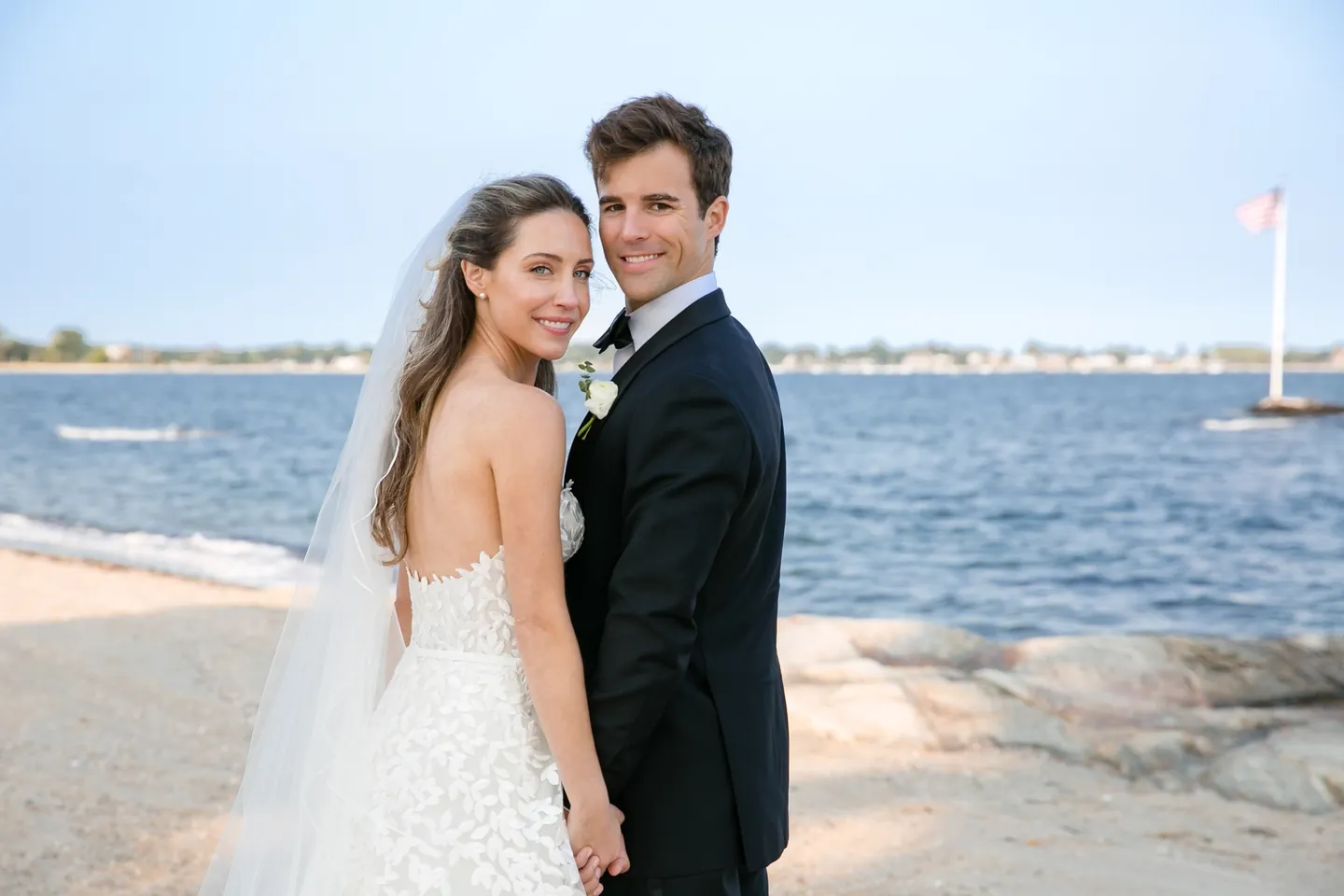 Britt and Andrew stand close on the sand with the blue water of the Sound behind