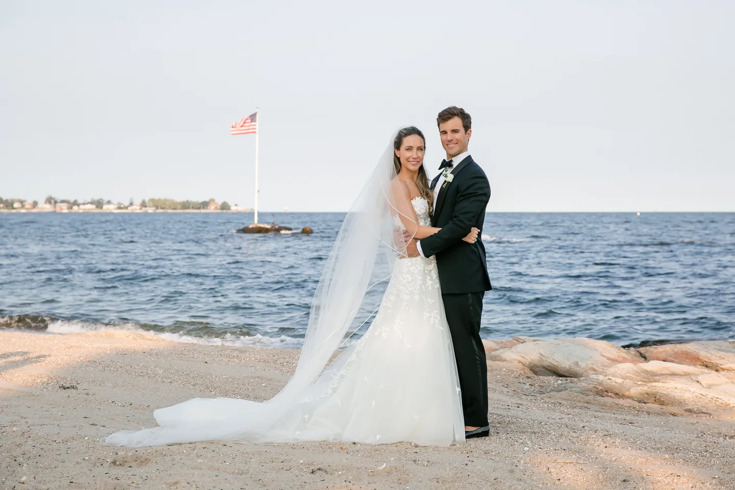 Britt and Andrew embrace on the beach, her long veil trailing across the sand toward the water