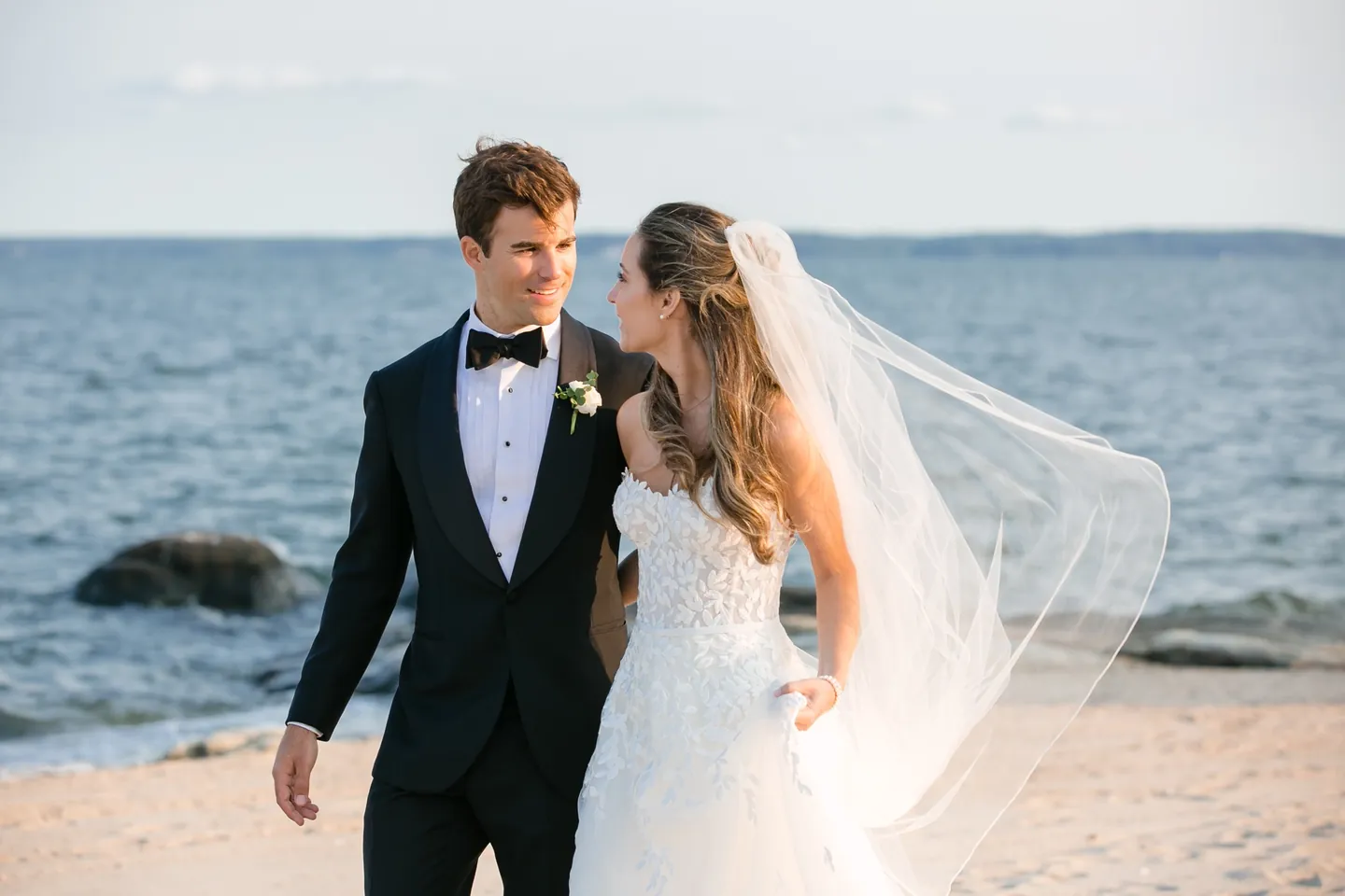 Andrew and Britt laugh walking along the waterline as her veil blows out behind her