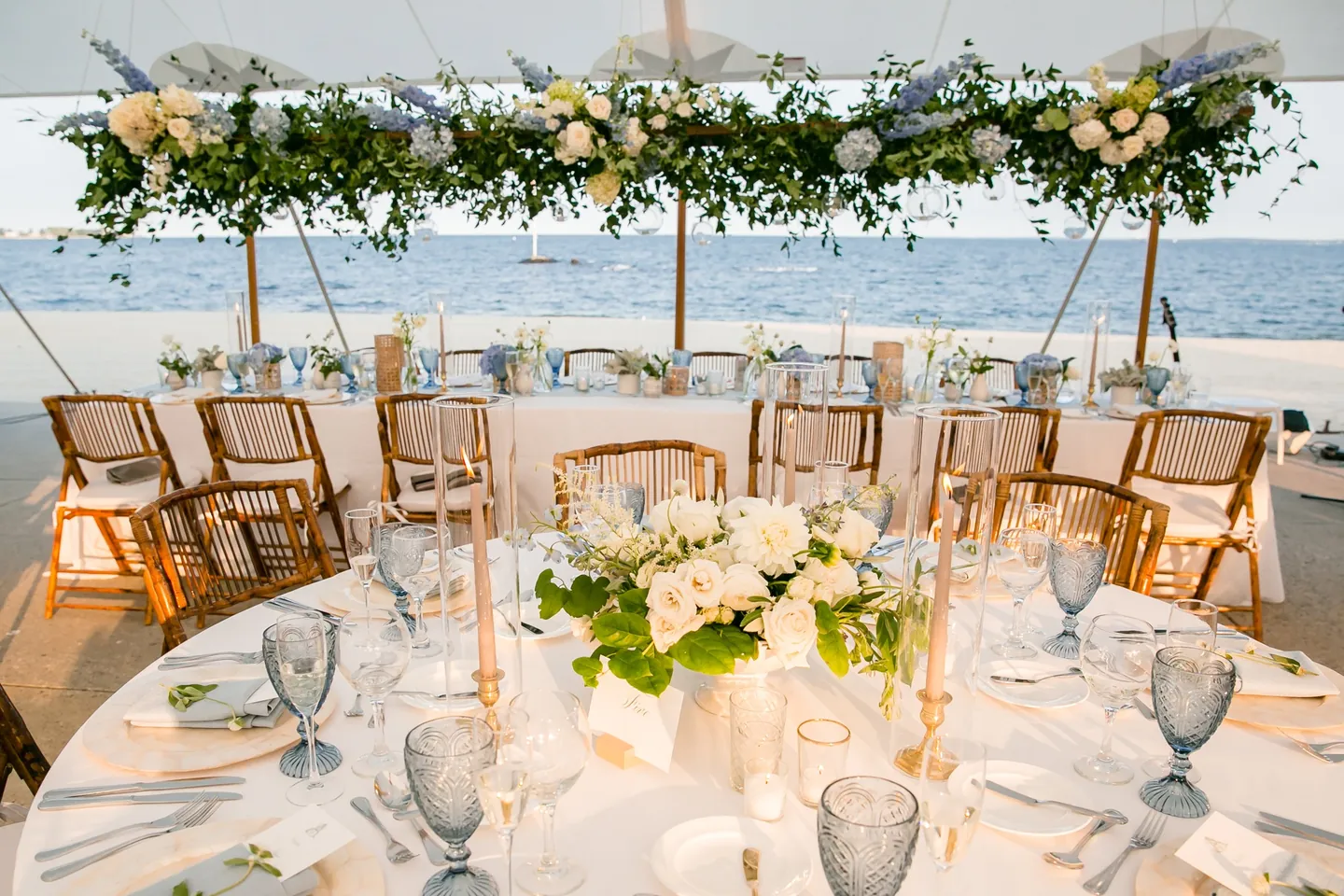 Bamboo chairs and round tables under a hanging garland of white and blue flowers facing the water
