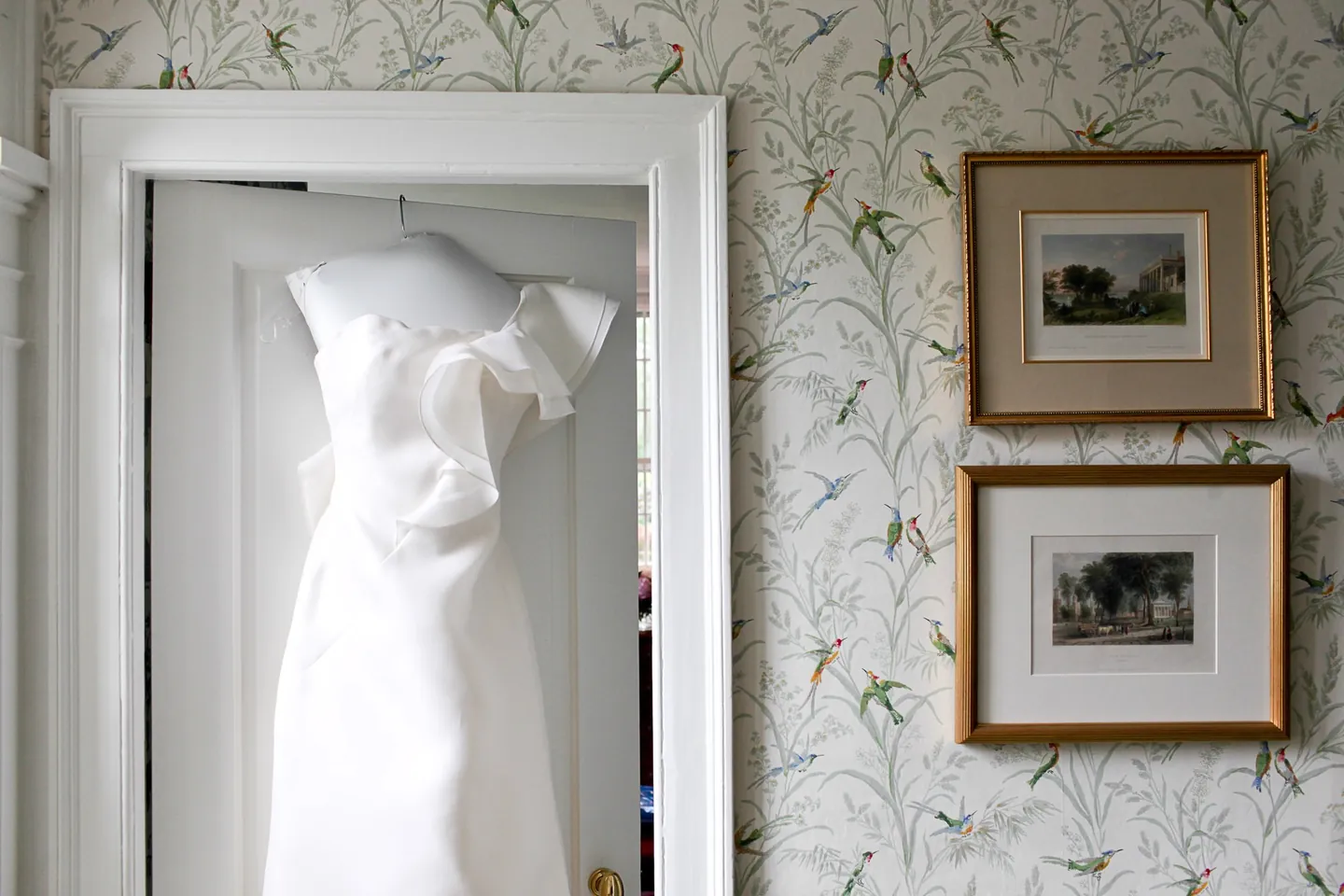 A white one-shoulder gown hangs on a door against green floral wallpaper