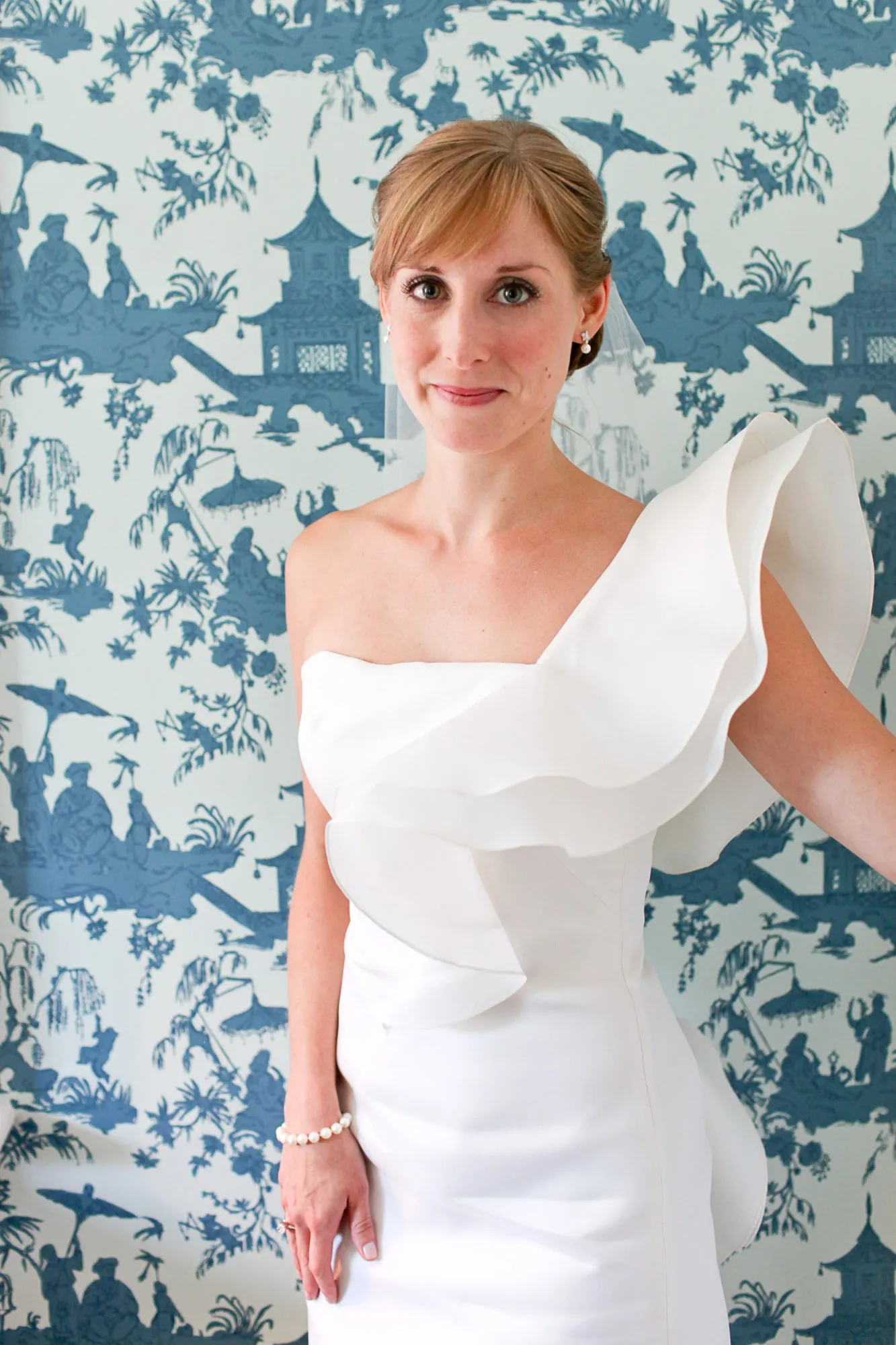 The bride in a sculptural one-shoulder gown poses against blue toile wallpaper