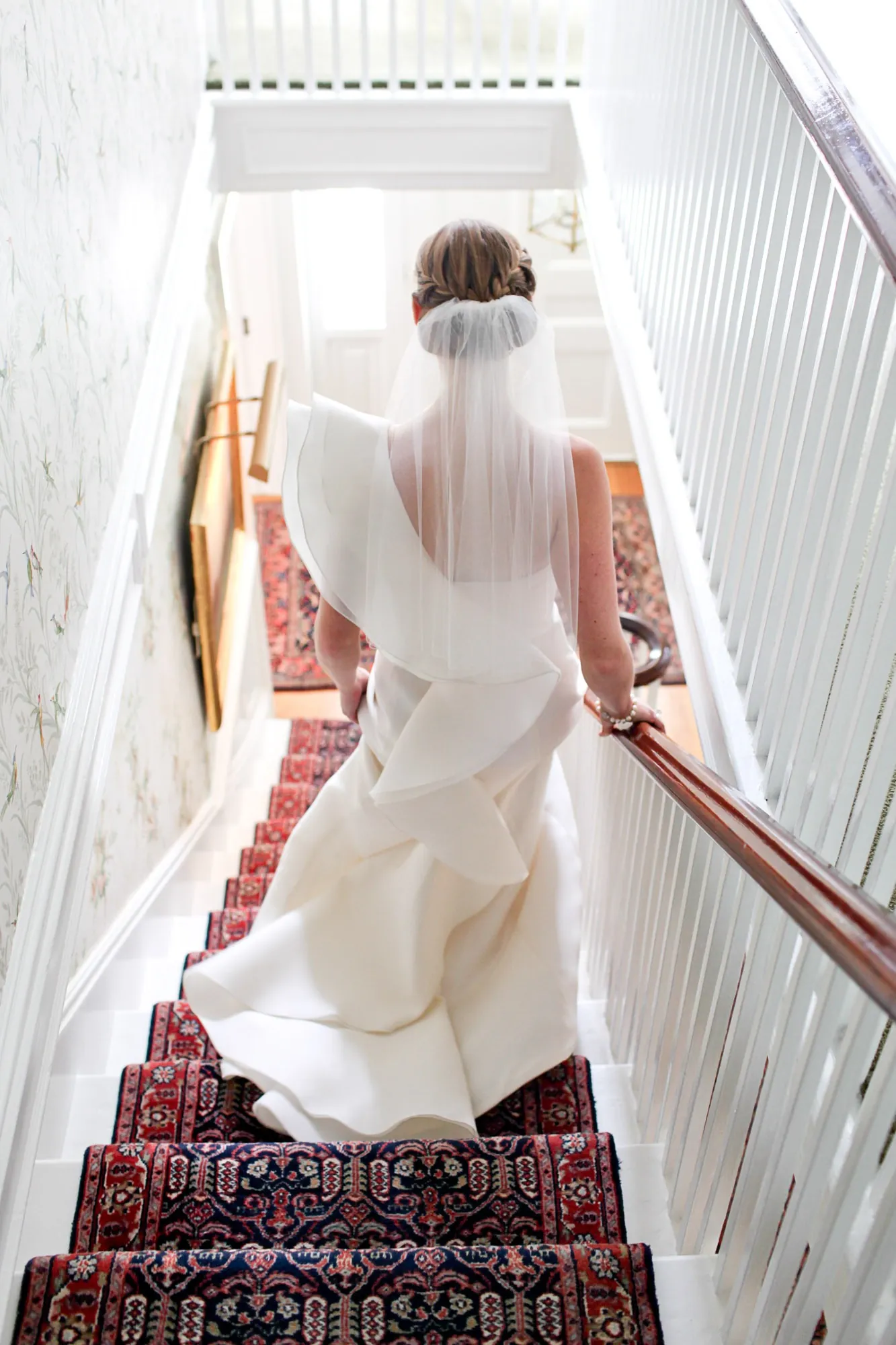 The bride descends a staircase with a red patterned runner, her veil trailing