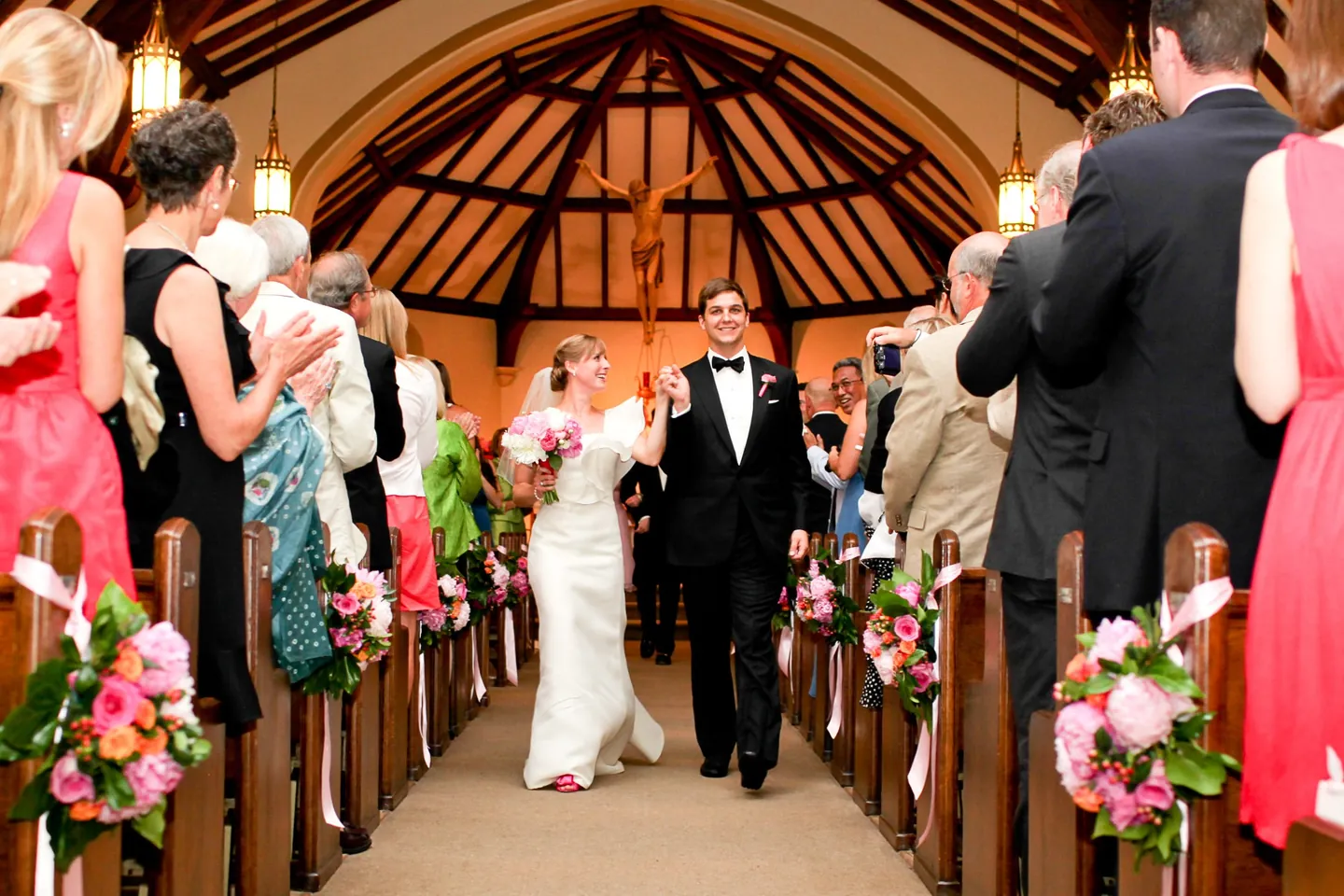 Caity and Matt walk up the church aisle under dark timber trusses and pink pew flowers