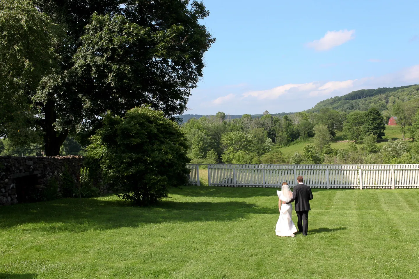 Caity and Matt walk across a wide lawn toward a picket fence and green hills