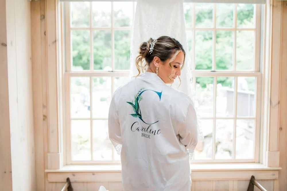 The bride in a white robe with her name on the back stands at a tall window
