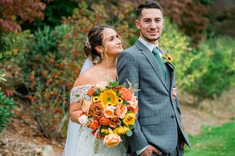 Caroline leans on Zachary's shoulder holding sunflowers, red fall foliage behind them