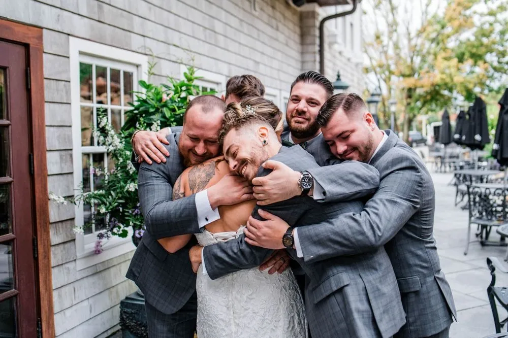 Groomsmen in gray suits wrap the bride in a big group hug outside the clubhouse