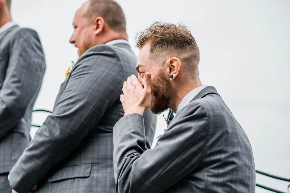 A bearded groomsman in a gray suit wipes tears from his eyes during the vows