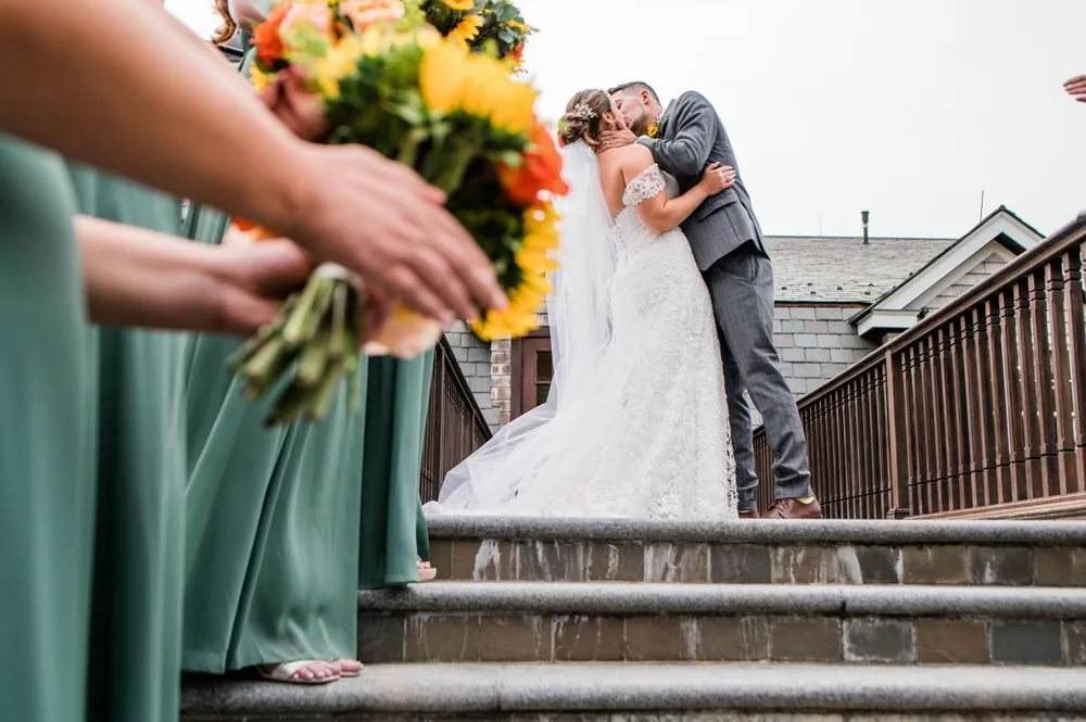 Caroline and Zachary kiss at the top of the stairs as bridesmaids hold sunflowers