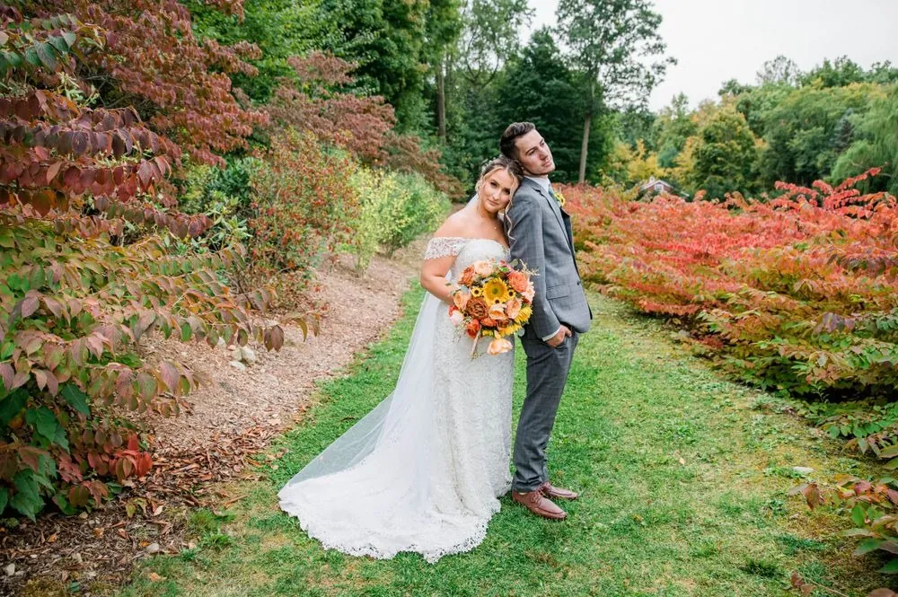 The couple stands close on a grassy path lined with red and orange fall shrubs
