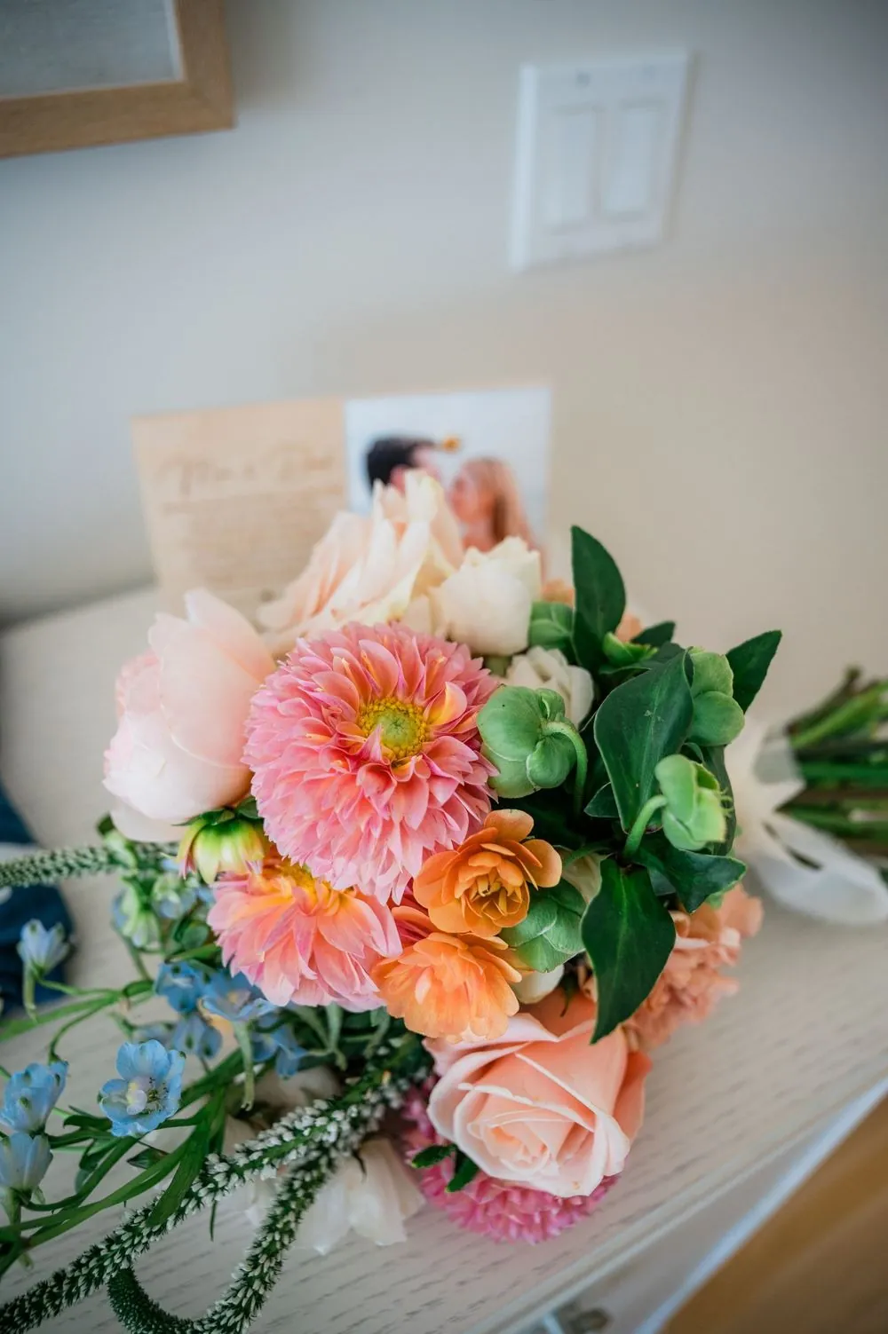Bouquet of coral dahlias, peach roses, and greenery lying beside a framed photo and note