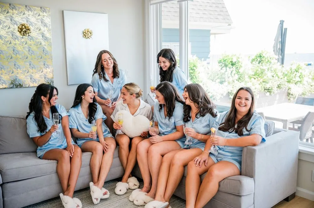 Bridesmaids in matching powder blue pajamas and slippers laugh together on a grey sofa