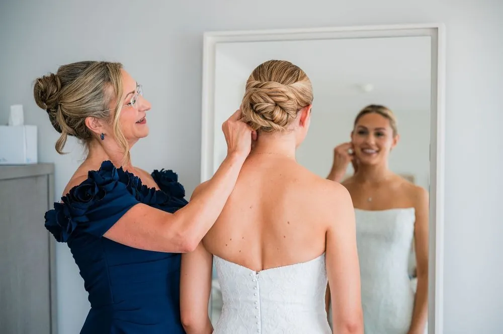 Woman in a navy dress pins the bride's braided updo while she watches in the mirror