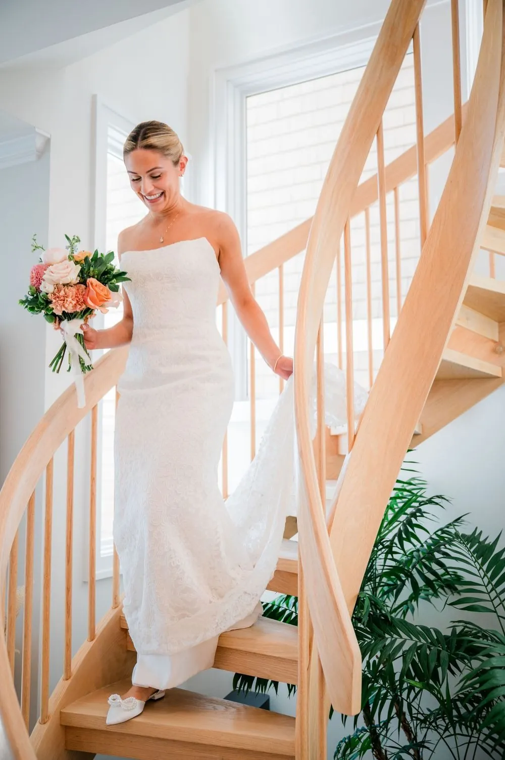 Bride walks down a curved wooden staircase holding her coral bouquet and lifting her skirt