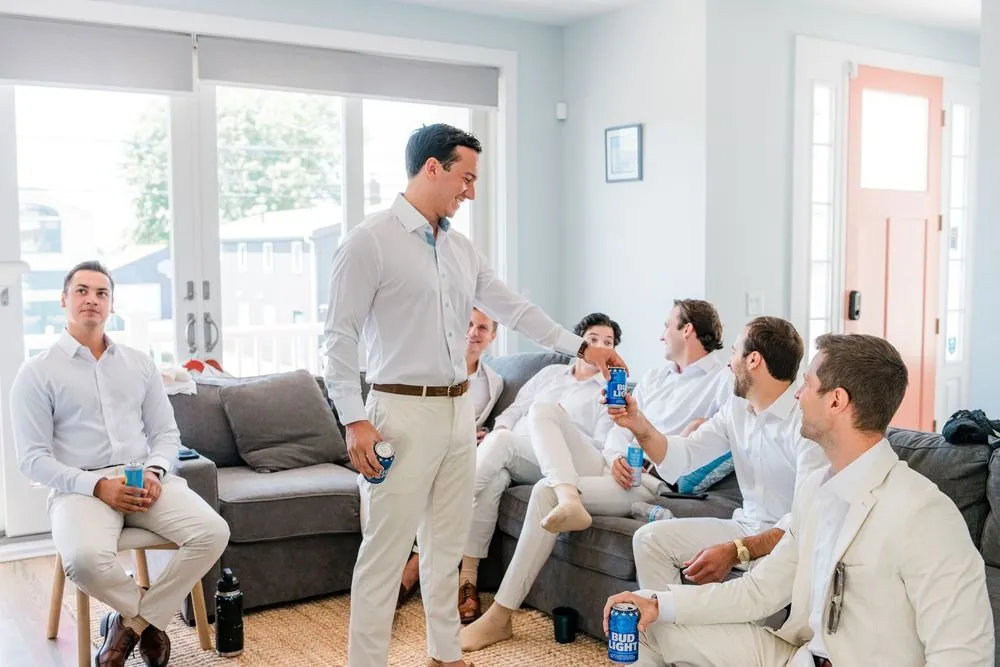 Groomsmen in cream trousers and white shirts relax on sofas with cans of beer