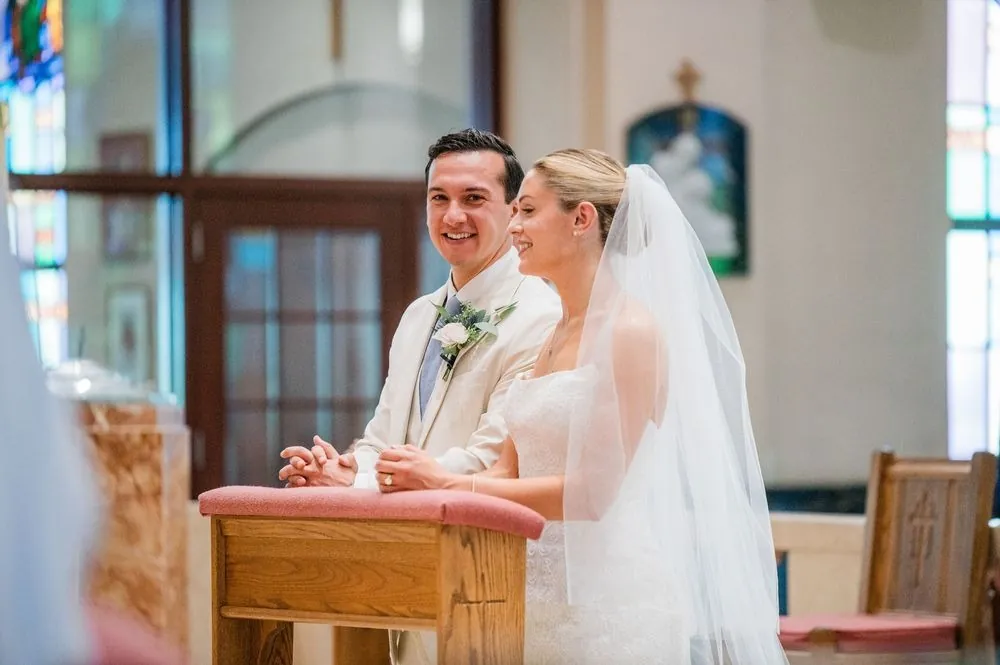 Chris and Carmen kneel at a wooden prayer bench, both grinning, her veil falling behind her