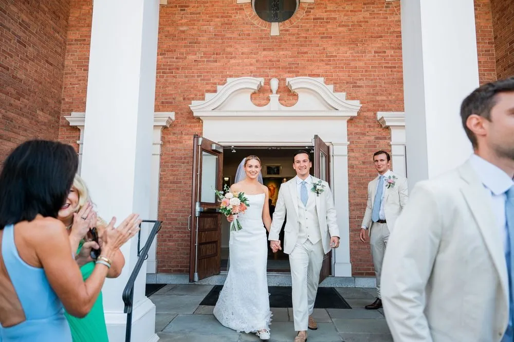 Chris and Carmen walk hand in hand out of a brick church doorway as guests clap