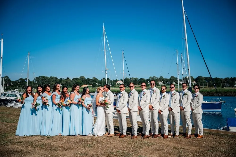 Wedding party in pale blue gowns and cream suits lined up on the grass at Southport Harbor