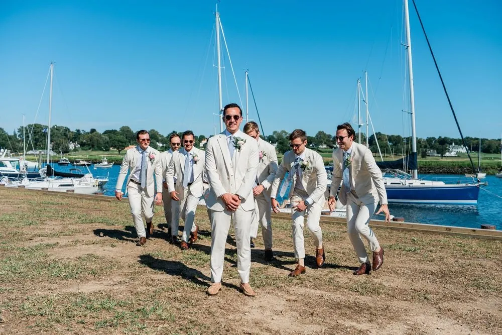 Groomsmen in cream suits and sunglasses stride across the harbor lawn with sailboats behind