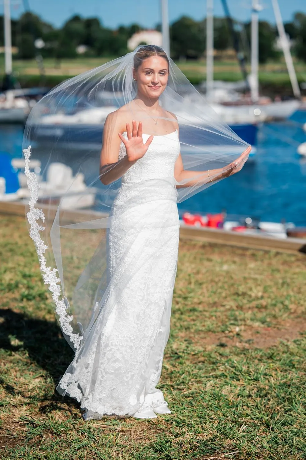 Bride lifts her long veil in the breeze on the harbor lawn with boats behind her
