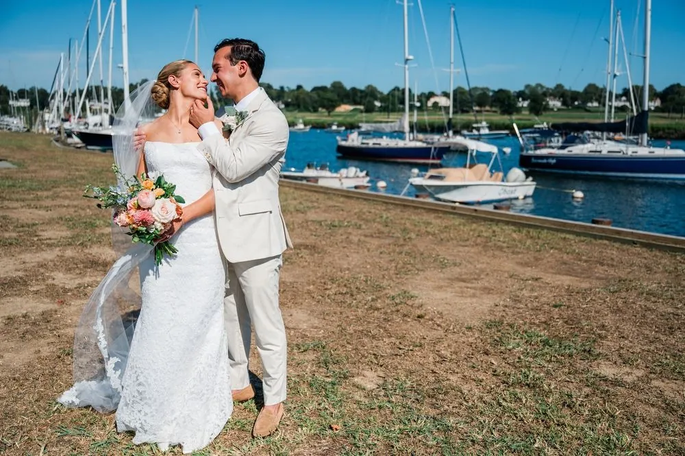Chris cups Carmen's face on the harbor lawn, sailboat masts and bright blue water behind
