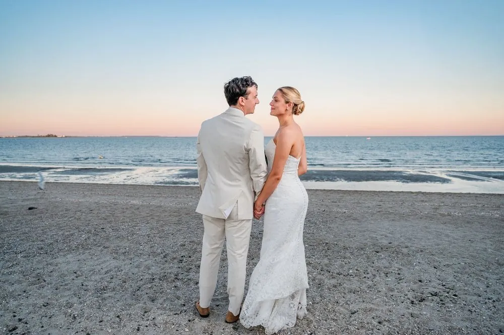 Chris and Carmen stand close on the beach facing the water as the sky turns pink at sunset