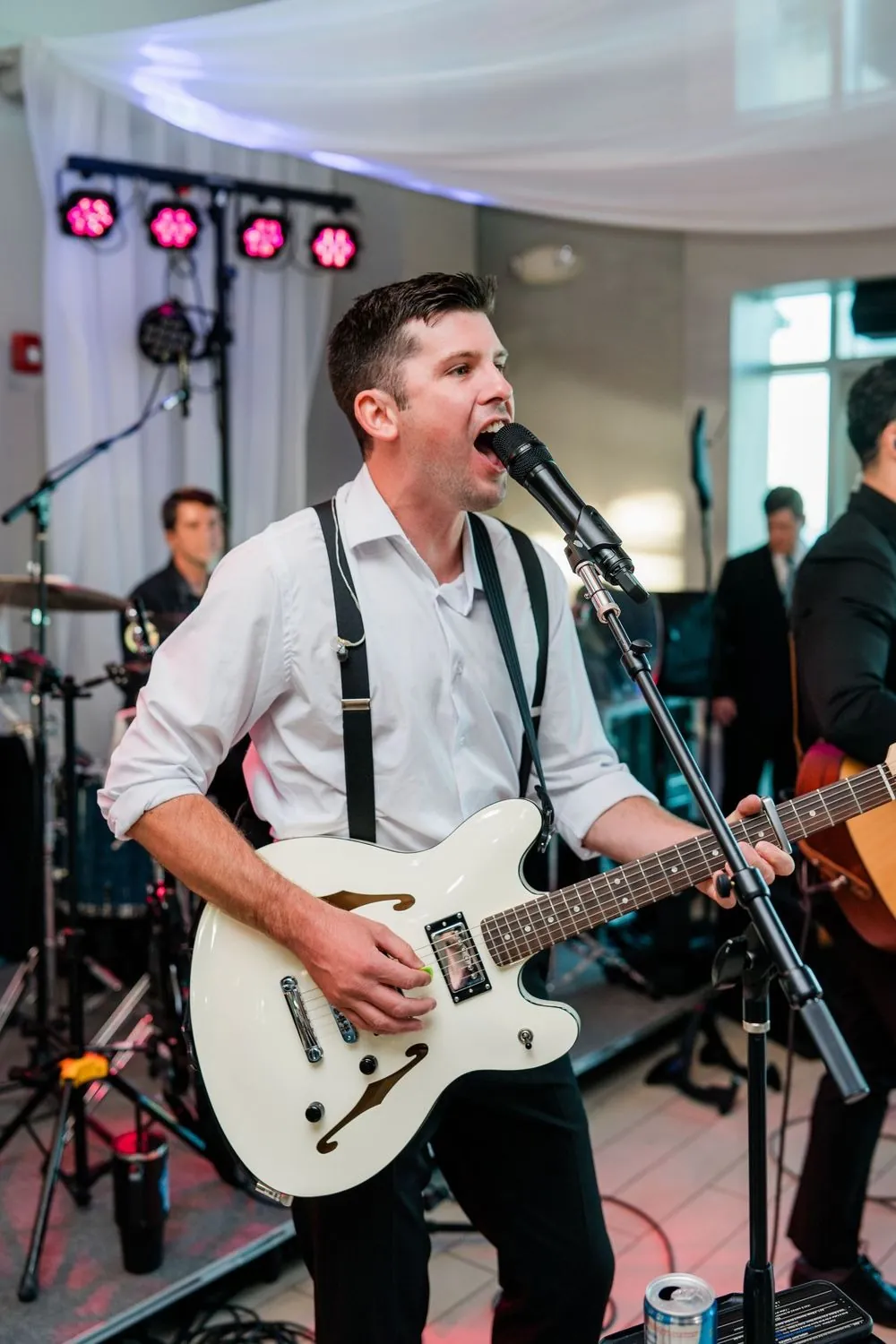 Band singer in suspenders plays a white electric guitar under pink stage lights
