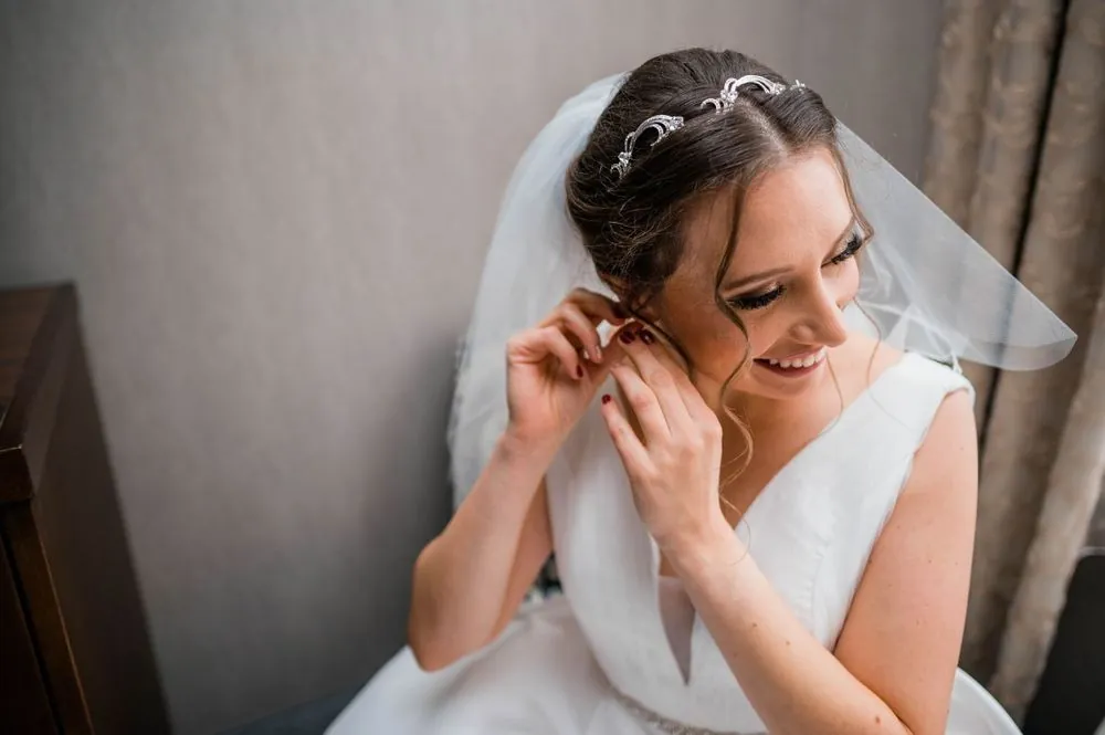 The bride smiles while putting on an earring, her veil pinned behind her