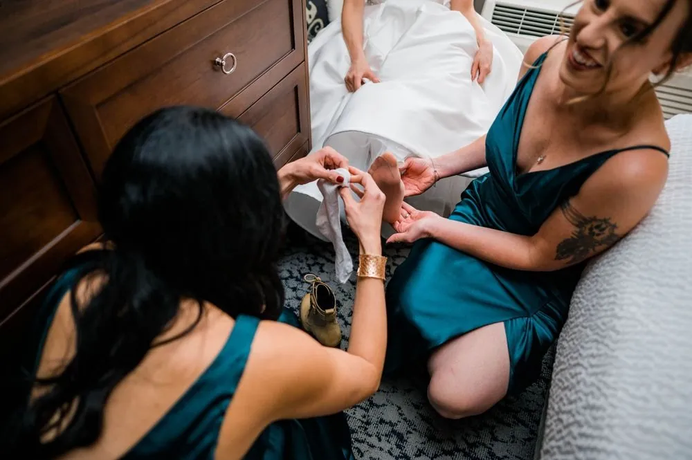 Bridesmaids in teal kneel to buckle the bride's shoes