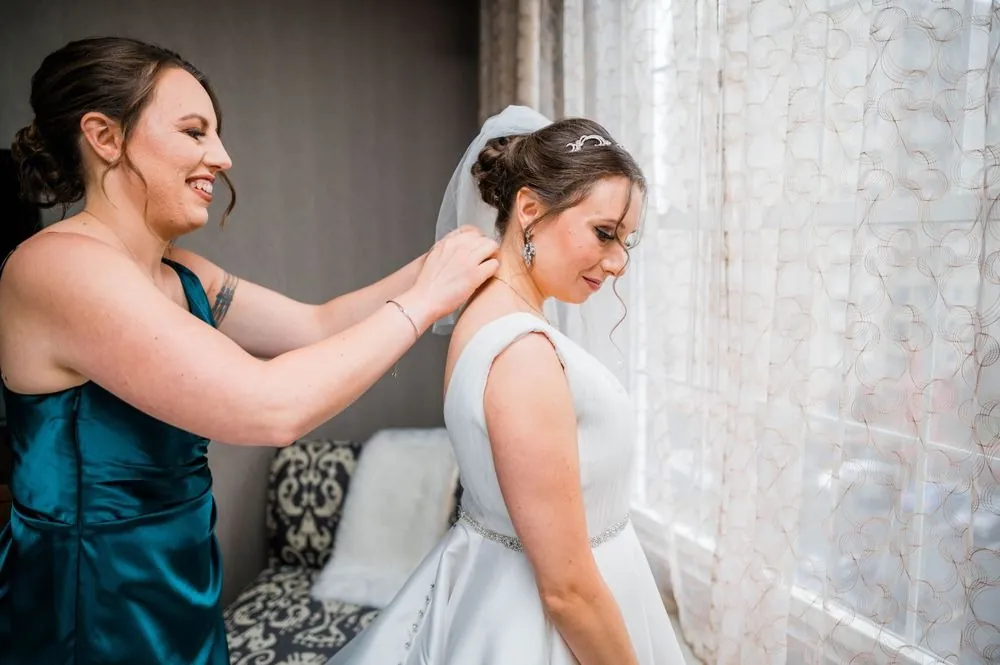 A bridesmaid in teal fastens the bride's necklace by a sheer-curtained window