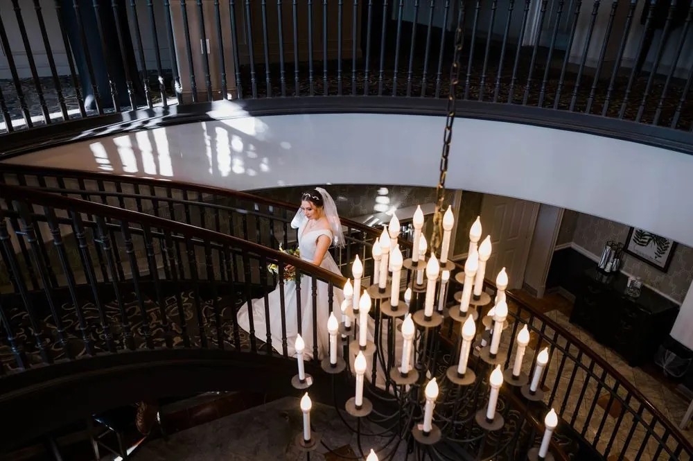 The bride walks down a curving iron staircase past a chandelier of tall white candles