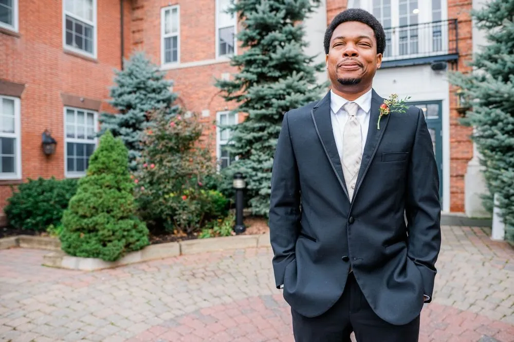 The groom in a black suit stands in a brick courtyard with evergreens behind him