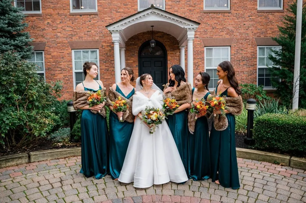 Bridesmaids in teal gowns and brown fur stoles surround the bride outside the brick building