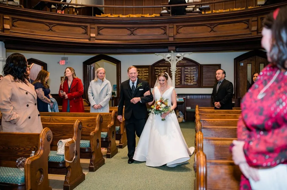The bride walks down the church aisle on a man's arm between wooden pews