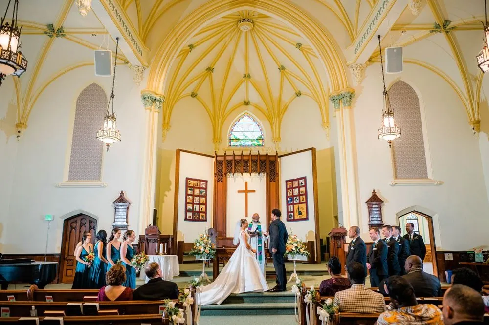 Wide view of the ceremony beneath a pale yellow ribbed vault and stained glass
