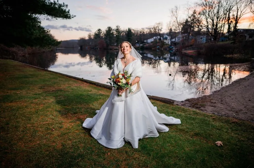 The bride stands on the lawn at Crystal Lake as the sunset glows on the water