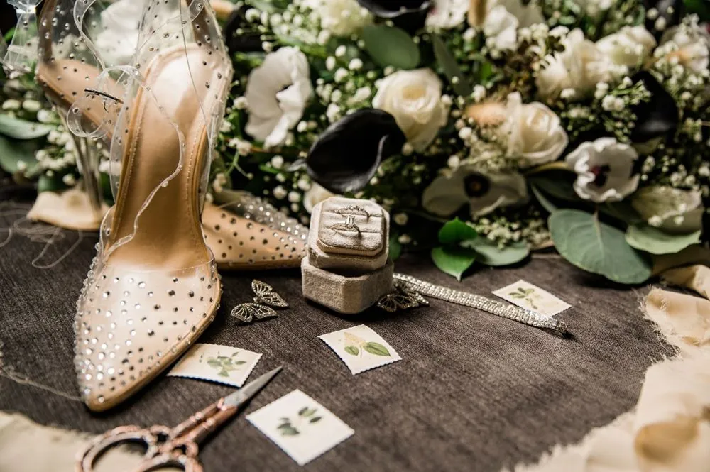 Sparkling clear pumps, a velvet ring box and pressed botanical stamps beside a white and green bouquet