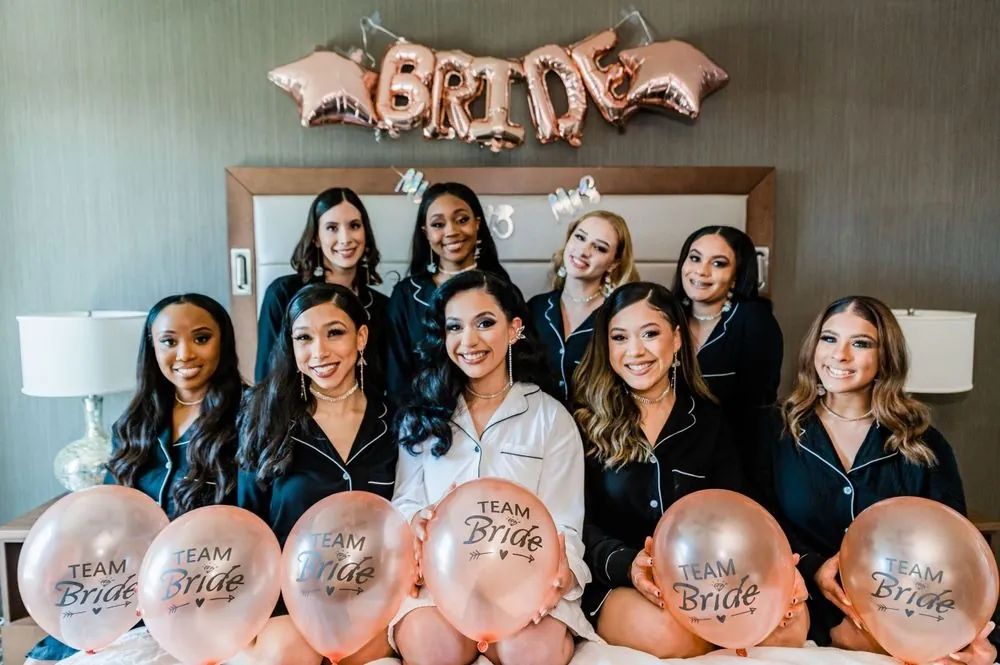 Bridesmaids in black pajamas hold Team Bride balloons around the bride on the bed