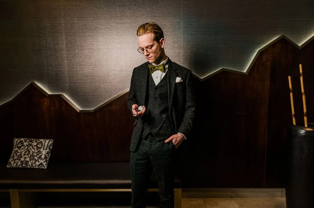 The groom in dark tweed and an olive bow tie checks his pocket watch against a lit wood wall