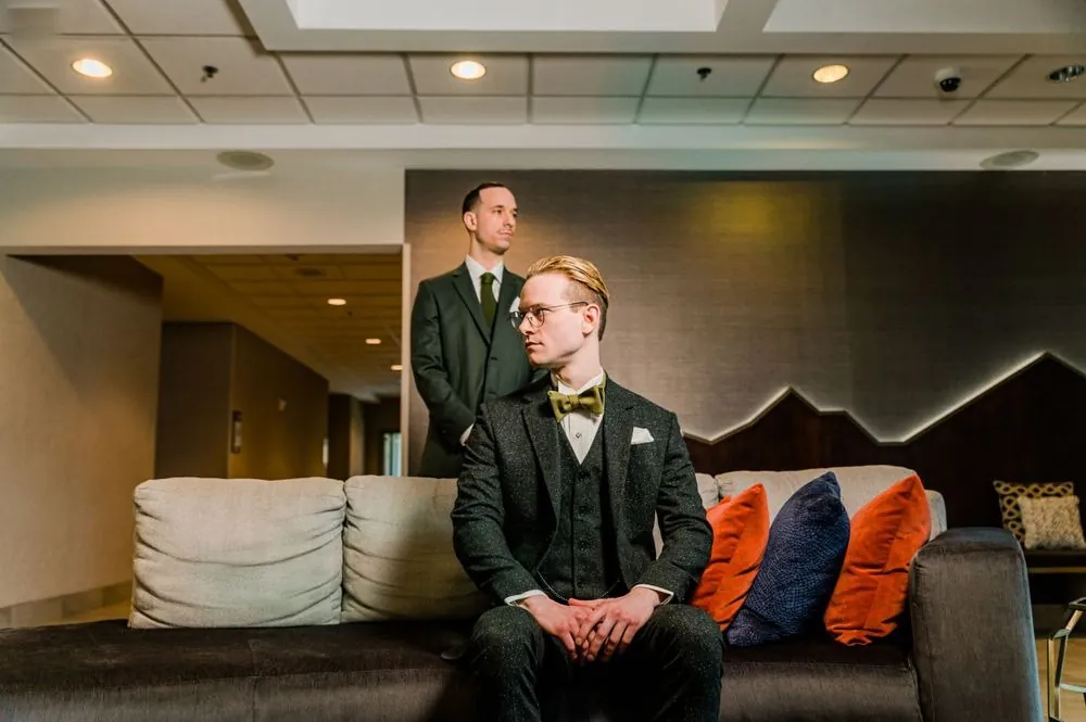 The groom sits on a gray sofa as a groomsman stands behind him