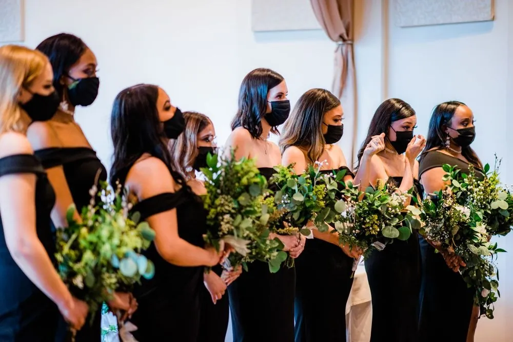Bridesmaids in black gowns and masks hold greenery bouquets together