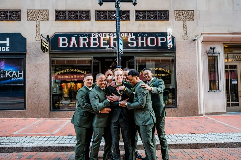 Groomsmen in green suits pile onto the groom outside a vintage barber shop