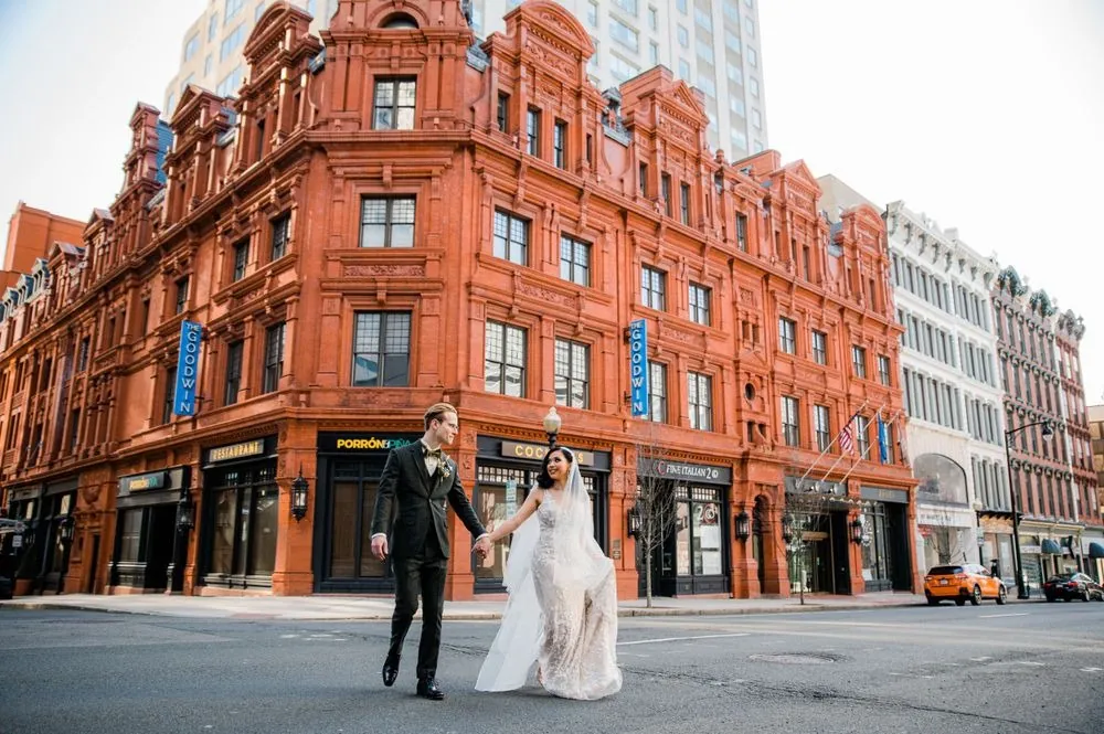 Victoria and Colin cross the street hand in hand before a red terracotta landmark building
