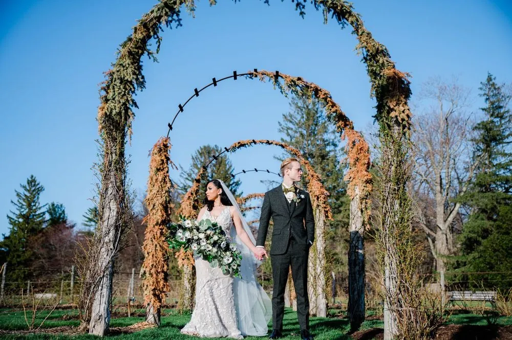 Victoria and Colin stand hand in hand beneath a line of vine-covered garden arches