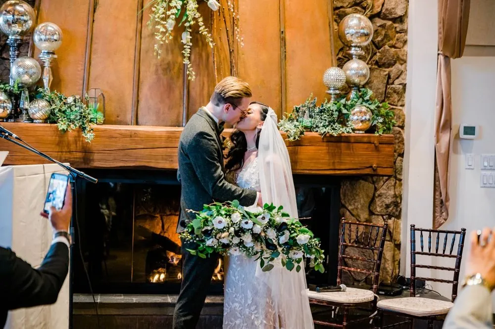 Victoria and Colin kiss in front of a lit stone fireplace as a guest films