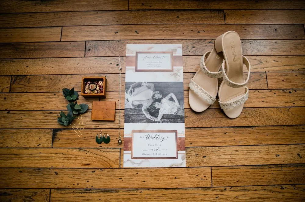 Invitation suite, lace sandals, green earrings, and a wooden ring box laid out on plank flooring