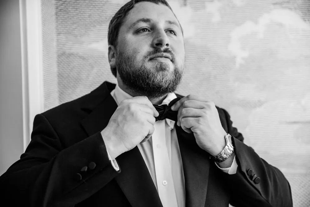 Black and white portrait of the groom adjusting his bow tie beside a bright window
